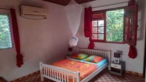 A bedroom with a white bed, red curtains, a window with a view of greenery, and an air conditioning unit on the wall.