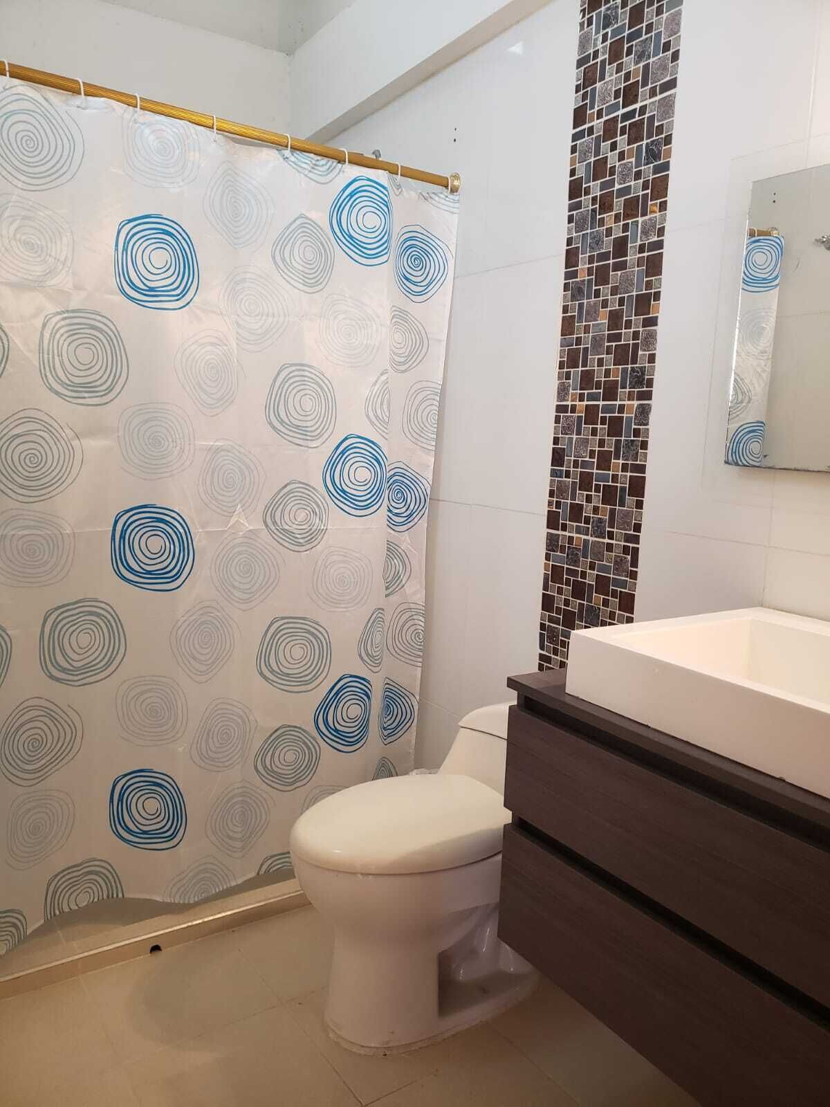 A bathroom with a toilet, a shower curtain with a geometric pattern, a tiled wall, and a sink.