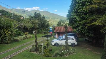 A white car parked in a lush green garden with a red-roofed building in the background.