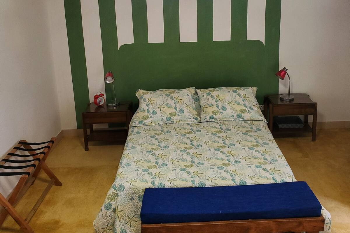 Un dormitorio con cama, un banco, dos mesitas de noche, una lámpara colgante y una ventana con vistas al follaje.