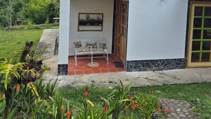 Edificio de dos pisos con un balcón, un pequeño patio con mesa y sillas, y un jardín con plantas tropicales.