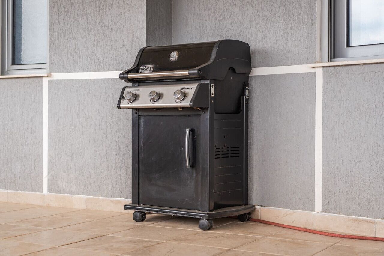 Una barbecue Weber Spirit portatile con quattro fuochi, un pensile laterale e uno sportello di vano portaoggetti anteriore, posizionata su un pavimento piastrellato di fronte a un muro grigio con finestre.