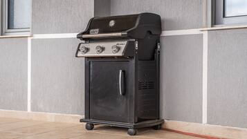 Una barbecue Weber Spirit portatile con quattro fuochi, un pensile laterale e uno sportello di vano portaoggetti anteriore, posizionata su un pavimento piastrellato di fronte a un muro grigio con finestre.