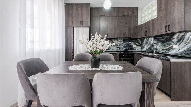 A modern kitchen with dark wood cabinets, a marble backsplash, and a dining table with grey upholstered chairs. - City Escape Apartment by PikHost (Shkodër)