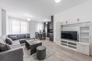 A modern living room with a grey sofa, a coffee table, and a TV on a white stand. There is a dining area with a table and chairs, and a kitchen in the background. - City Escape Apartment by PikHost (Shkodër)