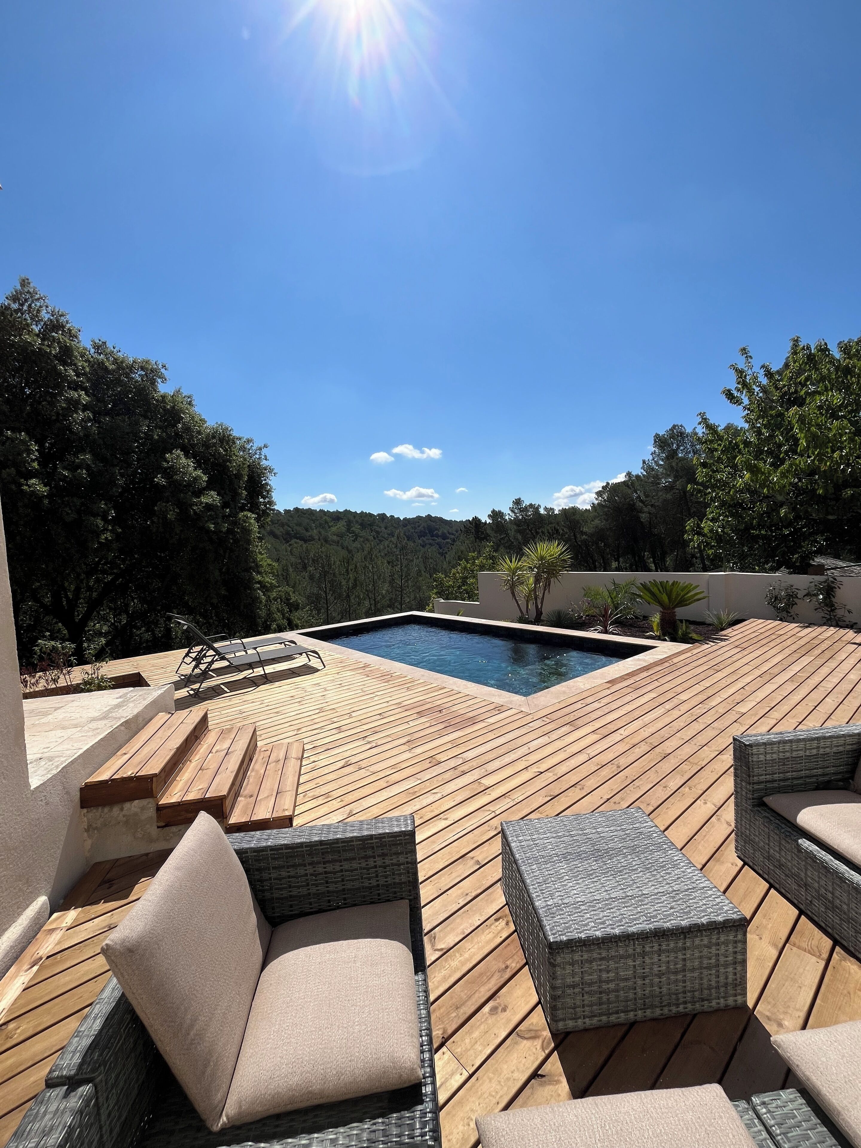 A wooden deck with a swimming pool, wicker furniture, and a view of a forested hillside.