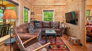 A cozy cabin interior with a wooden ceiling, a brown leather sofa, a small table, and a TV mounted on the wall. - The Three Dog Night Cabin (Bryson City)