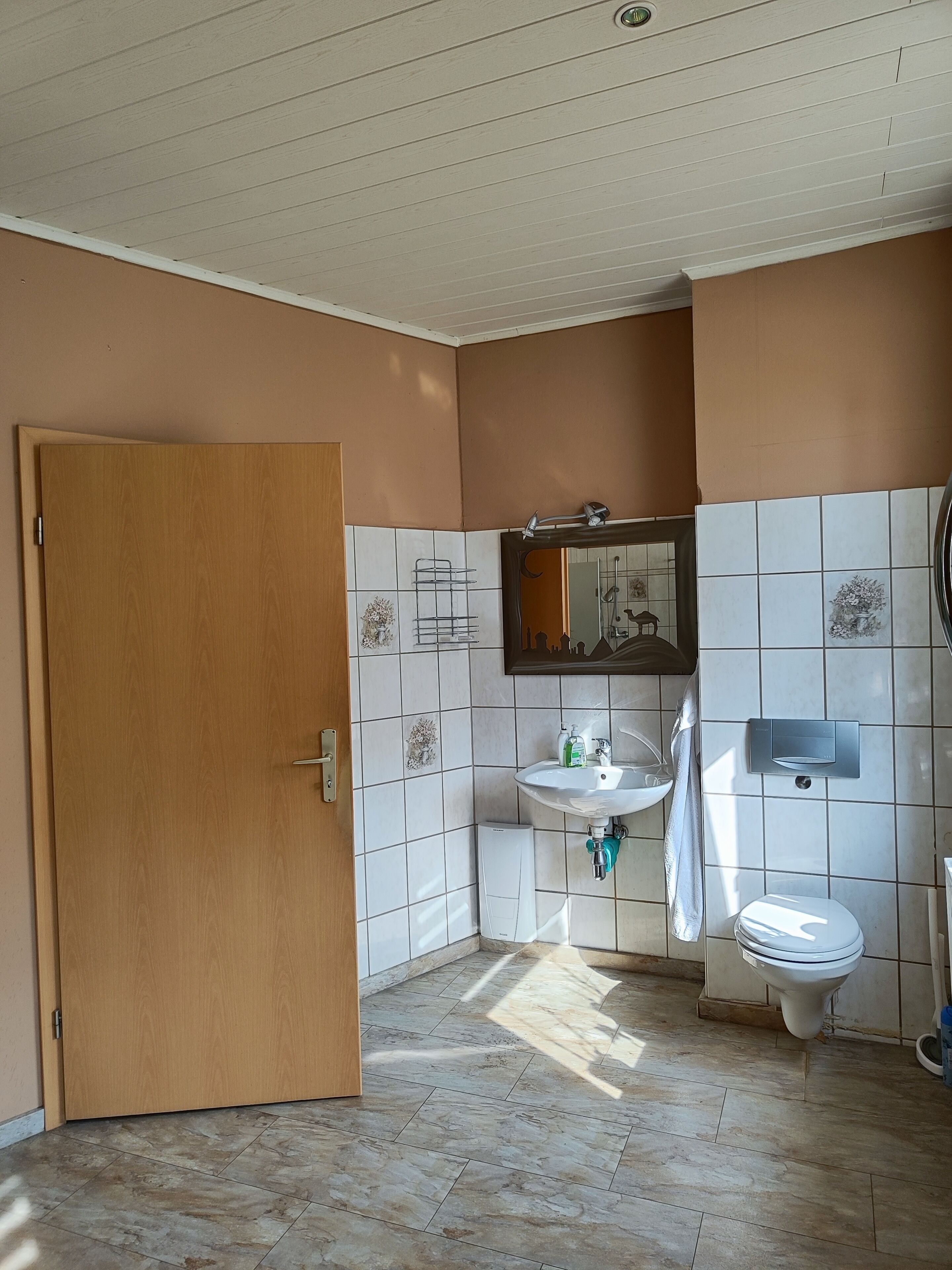 Une salle de bain avec une porte en bois, des murs recouverts de carreaux blancs, un lavabo, des toilettes et un miroir.