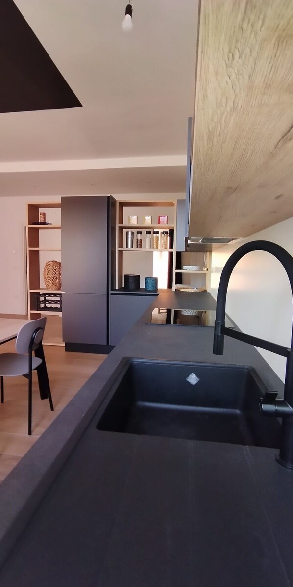 A modern kitchen with a built-in sink, a dining area with a chair, and a wooden shelf unit in the background.
