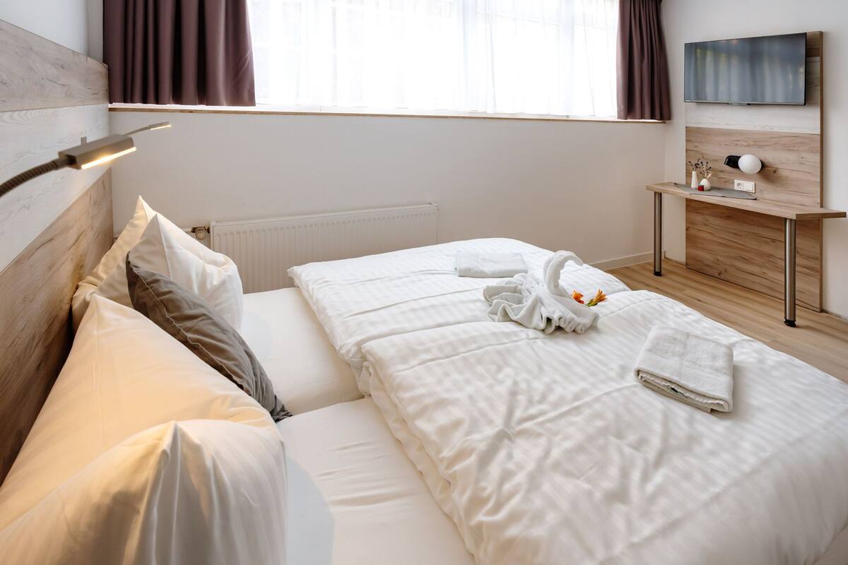 Une chambre d’hôtel avec un lit, une table de chevet avec une lampe, une télévision et une fenêtre avec des rideaux.