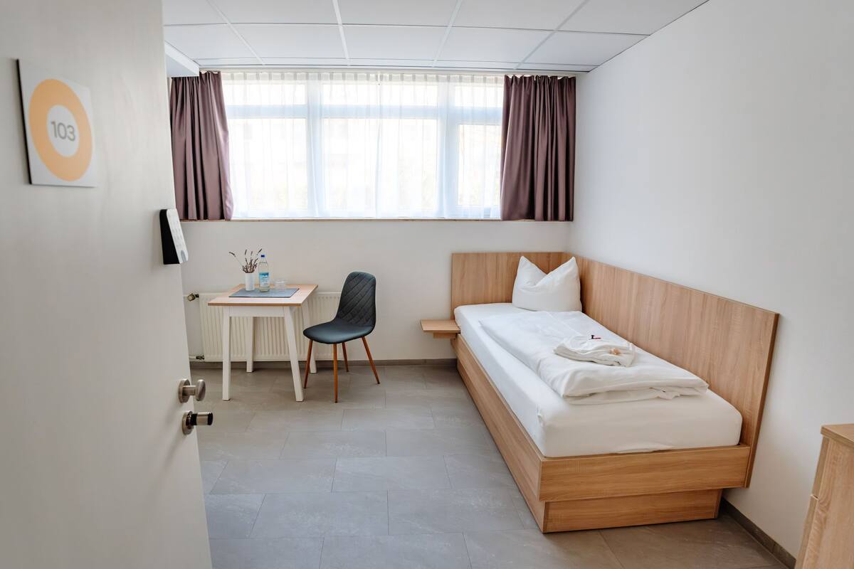Une chambre d’hôtel avec un lit en bois, un bureau avec une chaise et une fenêtre avec des rideaux.