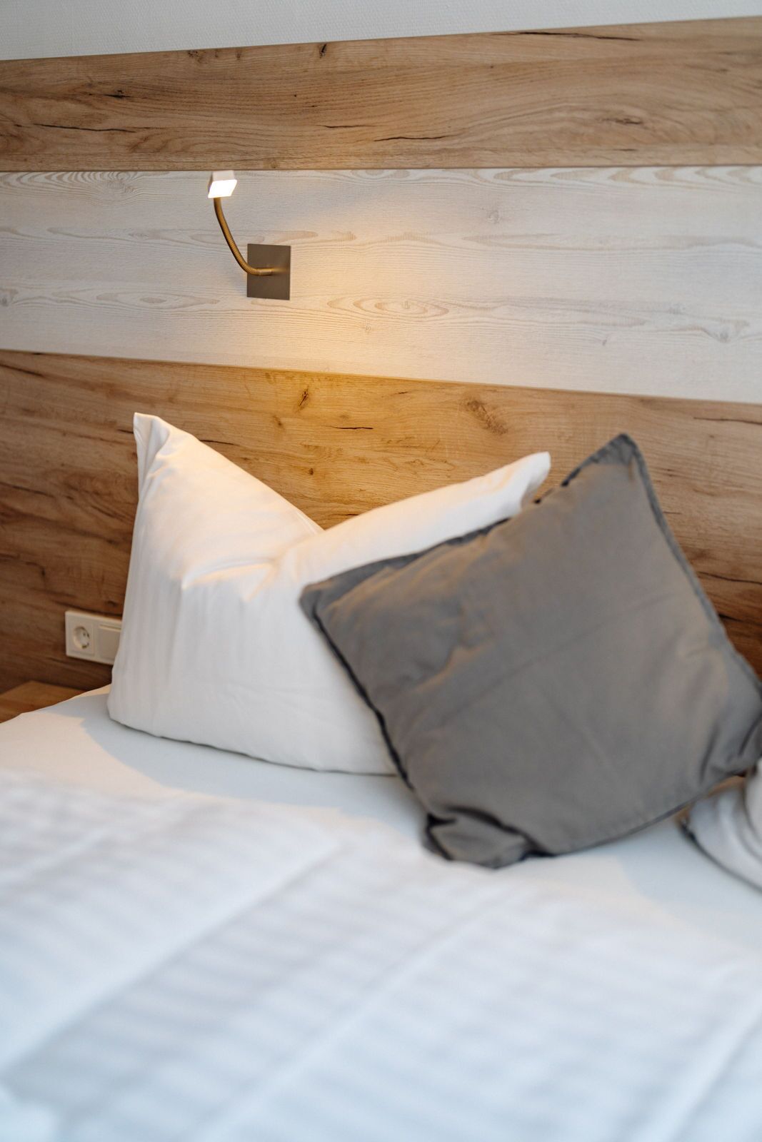 Un lit avec une literie blanche et deux oreillers, un blanc et un gris, adossé à une tête de lit en bois avec un interrupteur.