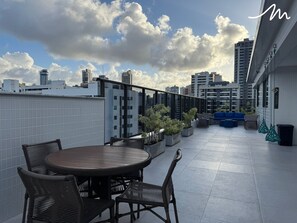 A rooftop terrace with a round table and chairs, potted plants, and a blue sofa. - COMPACTO TAMBAÚ (João Pessoa)