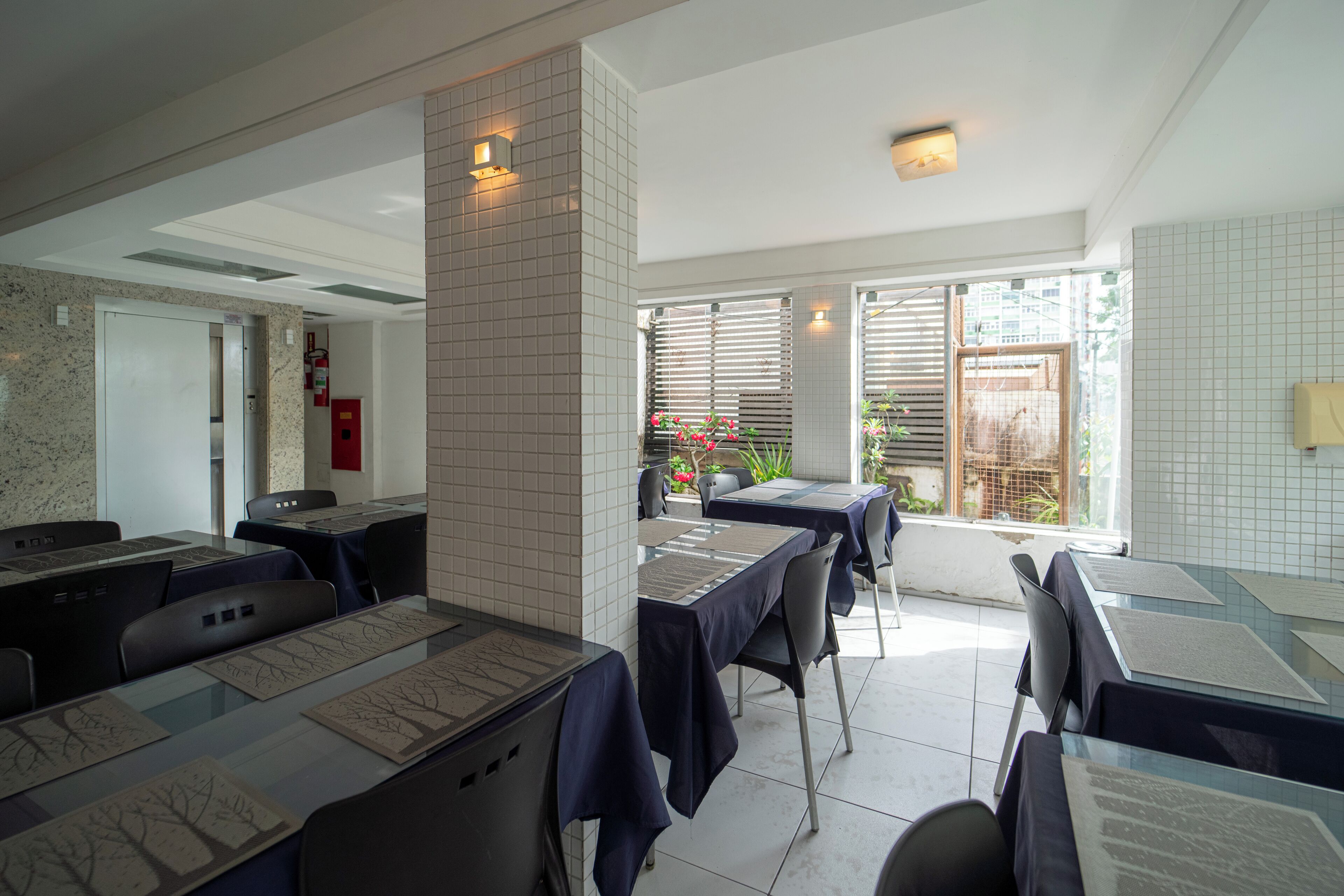Restaurante com mesas preparadas para refeições, parede com azulejos e janela com persianas.