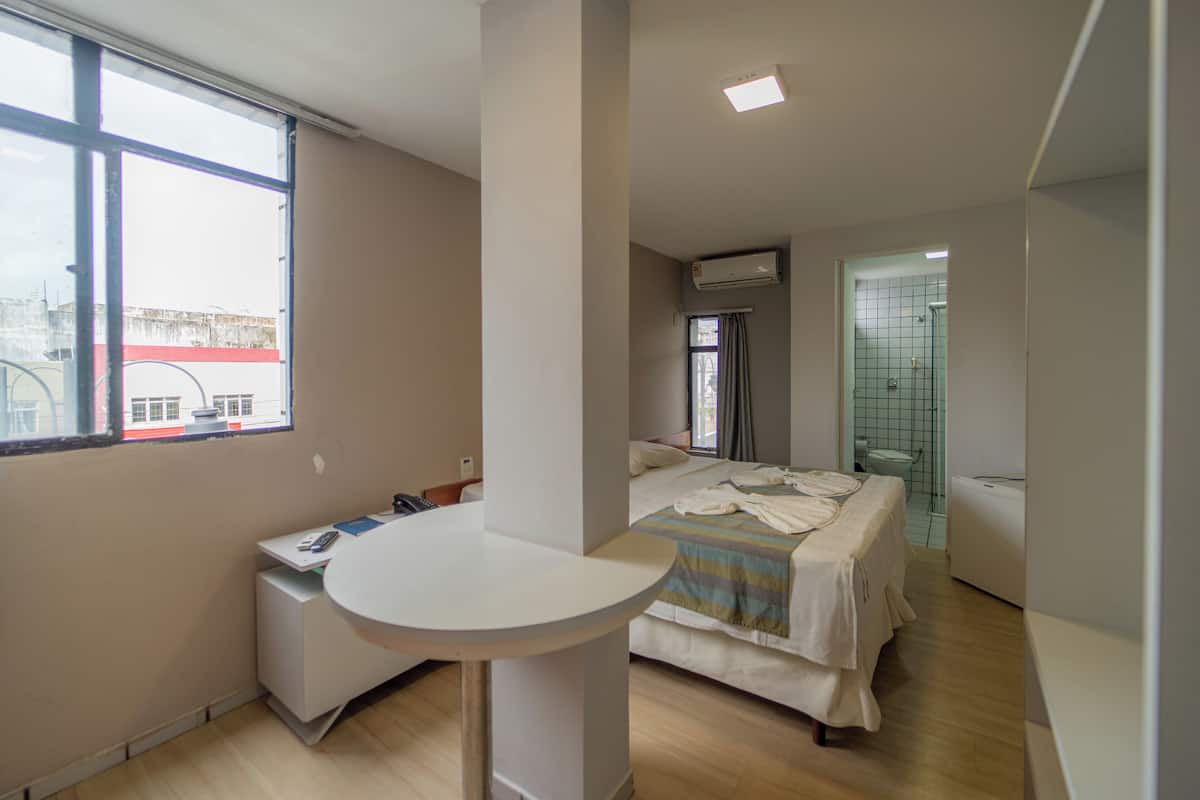 Een hotelkamer met een bed, een klein bureau met telefoon, een raam en een airconditioning.