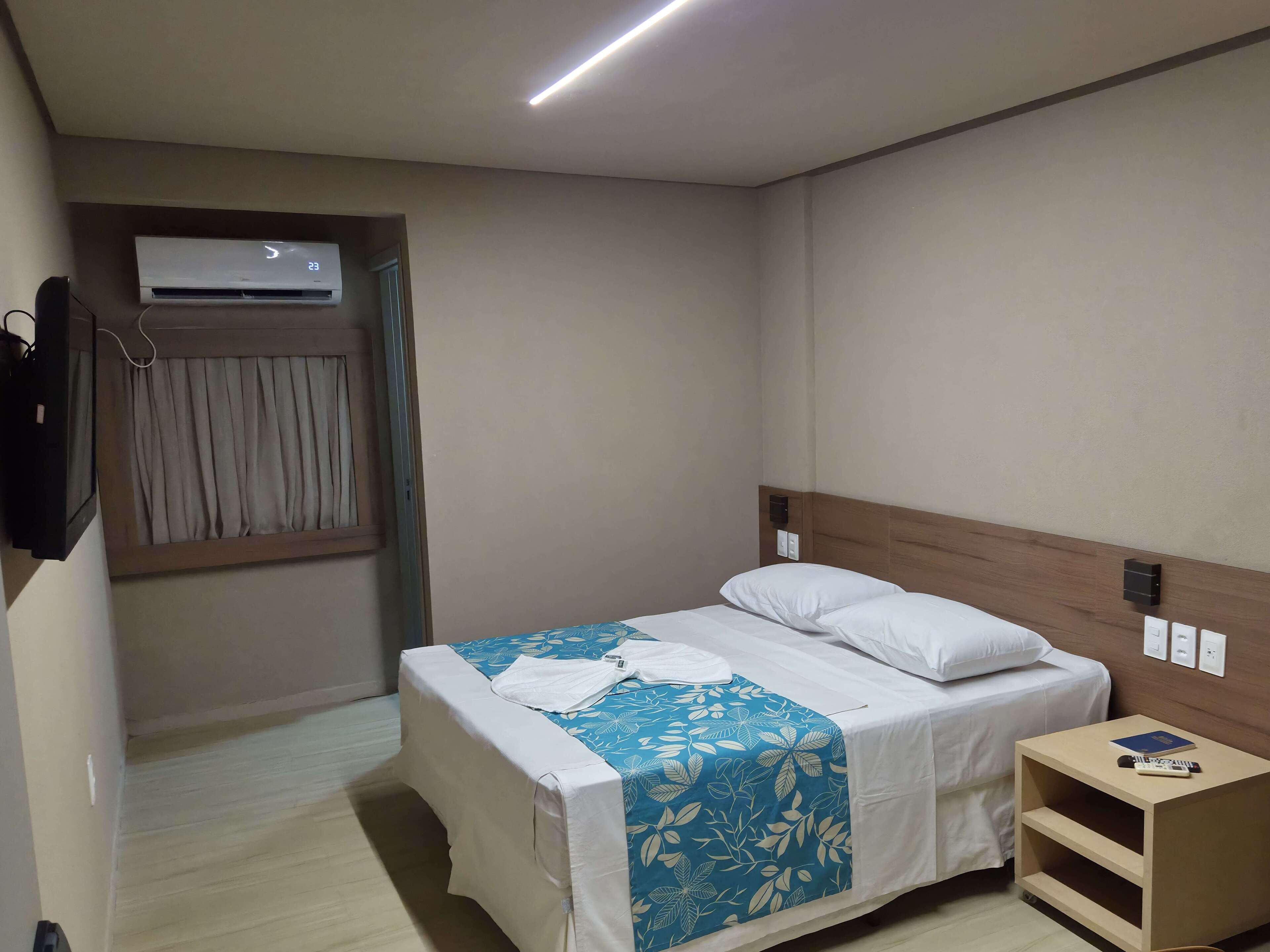 Una habitación de hotel con una cama, una mesita de noche con un libro y un control remoto, un equipo de aire acondicionado y una cortina.