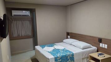 Una habitación de hotel con una cama, una mesita de noche con un libro y un control remoto, un equipo de aire acondicionado y una cortina.