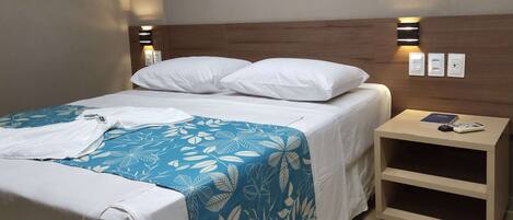 Una cama bien tendida con ropa blanca y una colcha de flores azules, una mesita de noche con un libro y un control remoto, y una lámpara de pared.