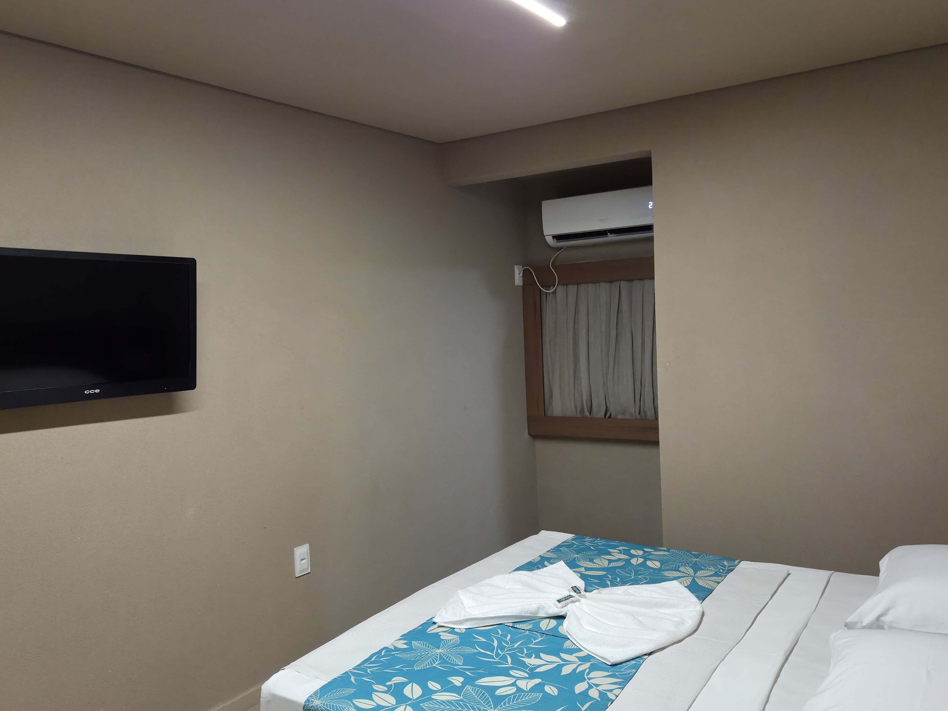 A hotel room with a bed, a flat-screen TV mounted on the wall, an air conditioning unit, and a window with curtains.