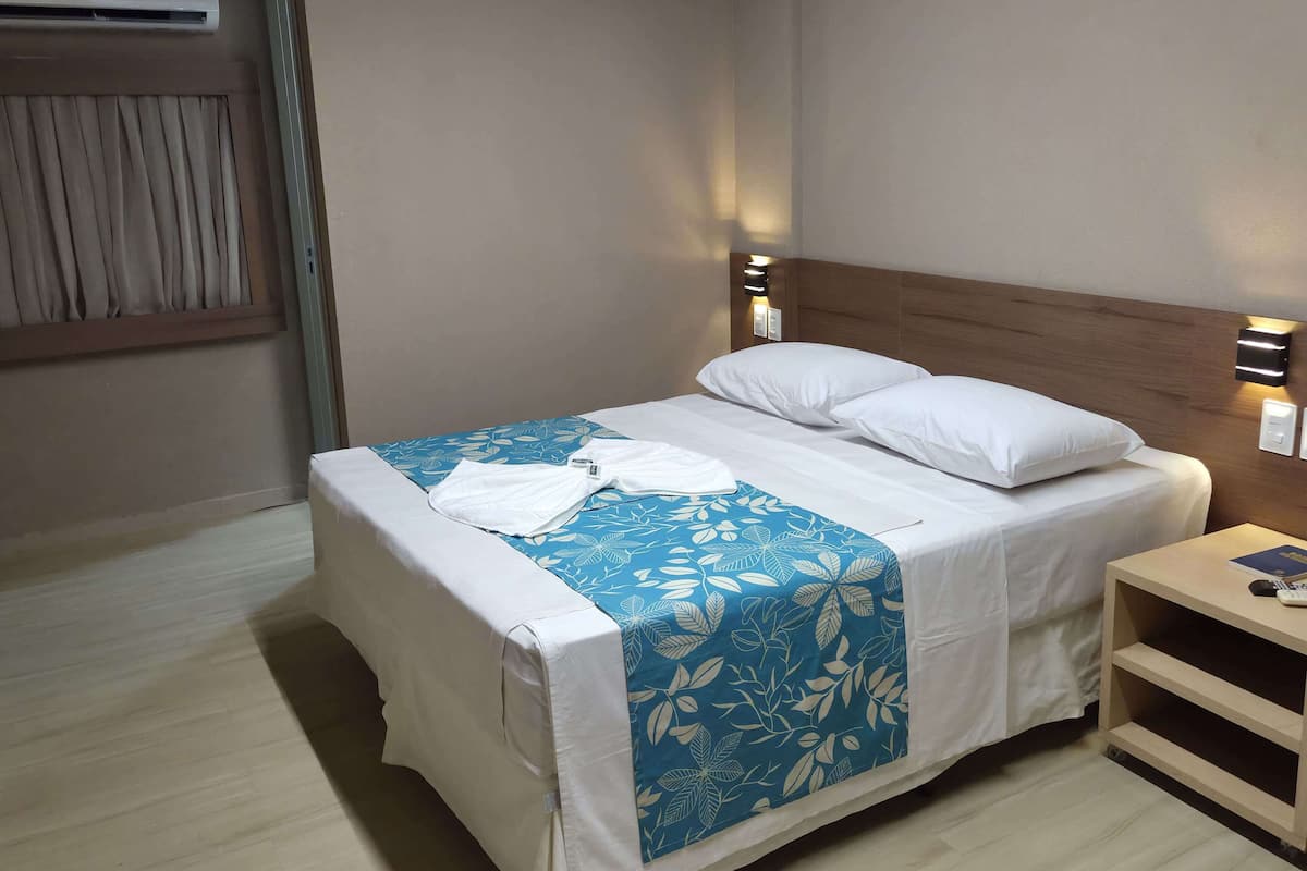 Een net opgemaakt bed met een blauwe bloemen spreideken, een wit kussen en een witte kussensloop. Er staat een houten nachtkastje met een boek en een afstandsbediening erop.