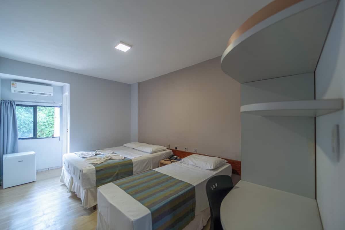 Een hotelkamer met twee bedden, een bureau, een stoel, een airconditioner en een raam met gordijnen.