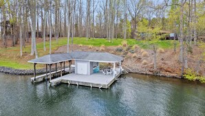 A floating structure with a covered deck, a small enclosed area, and a dock extending into a body of water, surrounded by trees. - Lakefront Lake Anna with Views, Dock, Fire Pit (Bumpass)