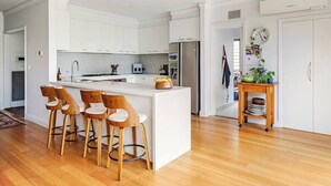 A modern kitchen with white cabinets, a central island with bar stools, a refrigerator, and a wooden side table with dishes. - Spacious Bayside Elwood Home. Walk to the beach, the botanical gardens, cafes.. (Elwood)