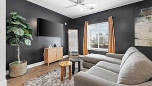 A modern living room with a grey sofa, a wooden cabinet, a large plant, and a flat-screen TV on the wall. - The Hidden Hills Suite The Gulch (Nashville)