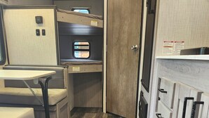 A compact RV kitchen with a wooden floor, a small dining table, and a kitchenette with a window. - Cozy RV Vacation (Antioch)