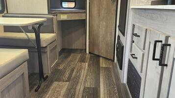 A compact RV kitchen with a wooden floor, a small dining table, and a kitchenette with a window.
