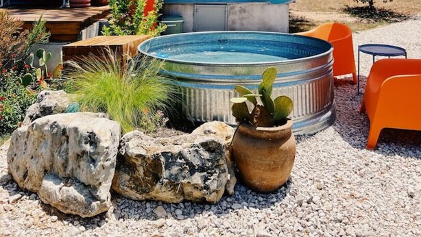 Una caravana azul con una pequeña ventana, una piscina circular y sillas naranjas.