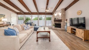 Un salon avec un canapé beige, une table basse, un téléviseur posé sur un meuble en bois et une cheminée en pierre.