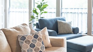 Un salon avec un canapé beige, un canapé bleu et un pouf assorti. Une grande fenêtre offre une vue sur la verdure à l’extérieur.