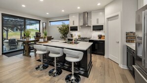 A modern kitchen with a central island, bar stools, and a large window overlooking a garden. - Elegant + Modern Home + Patio (Waukee)