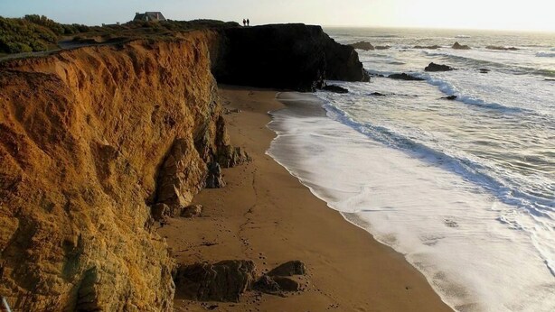 Uma praia costeira com um penhasco, ondas e uma estrutura distante.