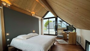 Quarto com cama grande, escrivaninha com computador e vista para o exterior através de uma grande janela.