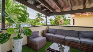 A covered outdoor seating area with wicker furniture and a glass table, surrounded by lush greenery and a view of buildings.