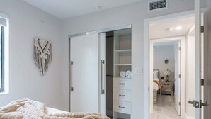 A bedroom with a bed, a large fluffy blanket, a macrame wall hanging, and a closet with open doors revealing a walk-in closet and a bathroom.