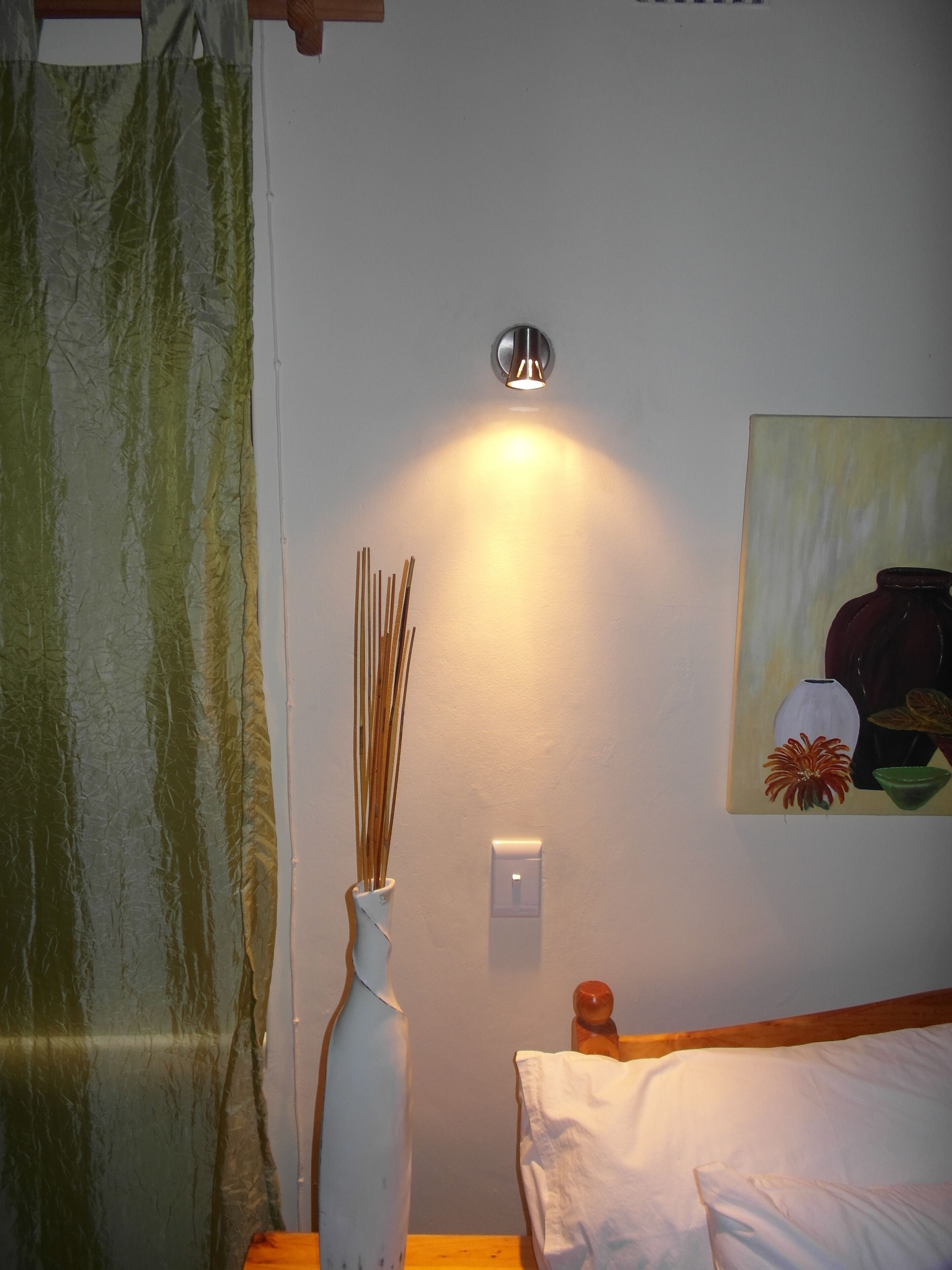 Una habitación con una cama, un jarrón blanco con ramas, un cuadro y una lámpara fijada a la pared.