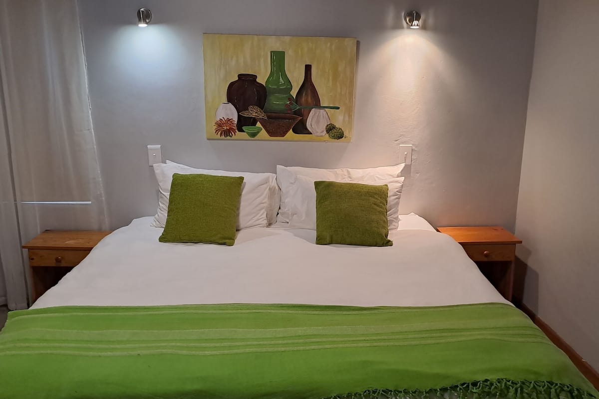 A neatly made bed with white and green bedding, two bedside tables, a painting on the wall, and a light fixture above the bed.