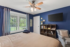 Um quarto com cama, ventilador de teto, estante, cadeira e janela com cortinas.