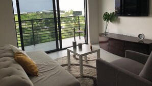 A modern living room with a large window, a sofa, a coffee table, and a TV mounted on the wall. - Modern building, centrally-located condo,2 bedrooms/2 bathrooms, parking space.  (Doral)