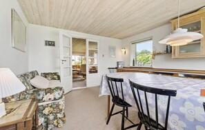 Un'accogliente stanza con soffitto in legno, un divano floreale, un tavolo da pranzo con sedie e un angolo cottura con pensili in legno e una lampada a sospensione.