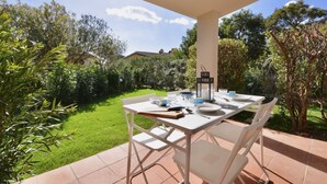 An outdoor patio with a table set for dining, surrounded by lush greenery and a clear sky.
