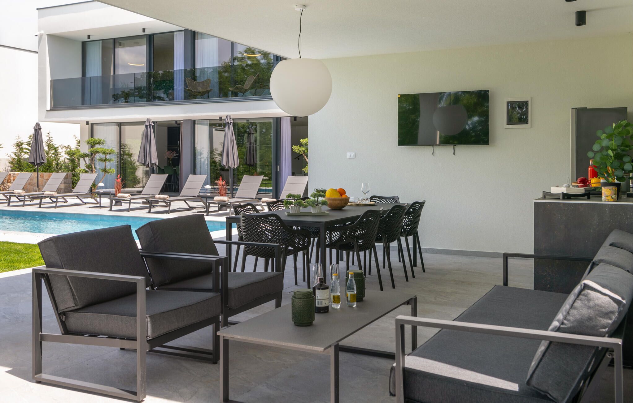 A modern outdoor living area with a pool, lounge chairs, a dining table, and a TV mounted on the wall.