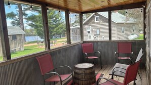 A covered outdoor seating area with wooden flooring, metal chairs, a wooden barrel, and a view of a lake and cabins. - Log Cabin on Lake Alice & ATV/UTV Trails, Sister Bear 2-Bedroom (Tomahawk)