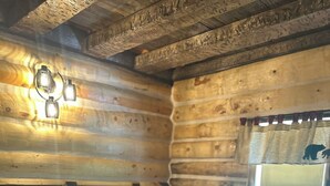 A log cabin room with a bed, a headboard, a window with a bear silhouette curtain, and a wall-mounted light fixture. - Log Cabin on Lake Alice & ATV/UTV Trails, Sister Bear 2-Bedroom (Tomahawk)