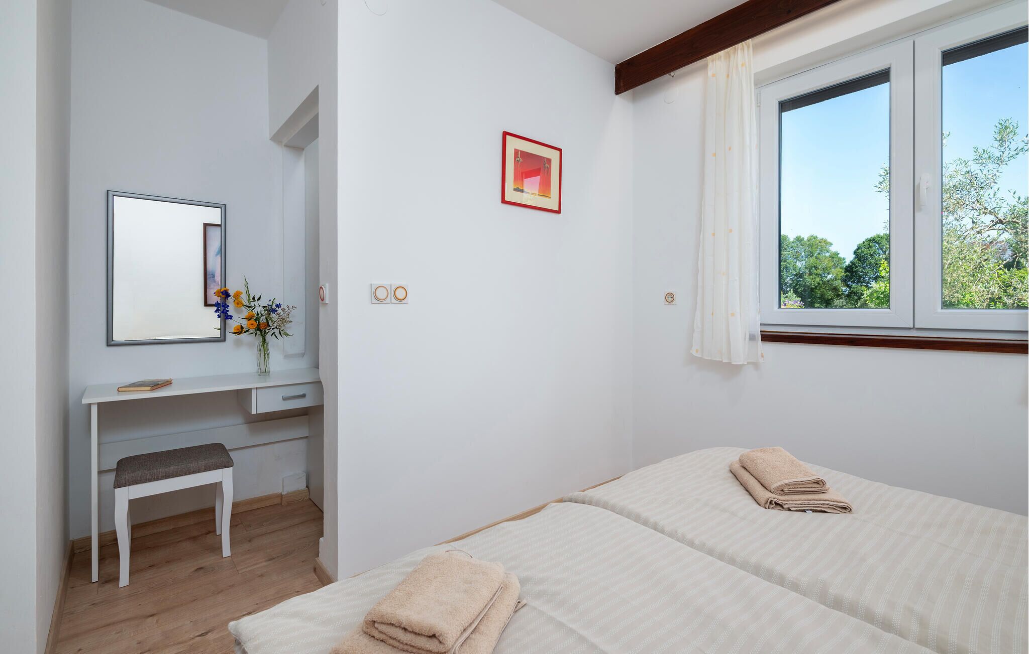 A bedroom with two beds, a desk with a mirror, and a window with a view of trees.