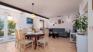 A dining area with a round table, wooden chairs, and a vase of flowers. There is a grey sofa and a small table with a plant in the living area.