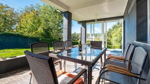 An outdoor patio with a glass table and wicker chairs, surrounded by a green hedge and a view of hills.